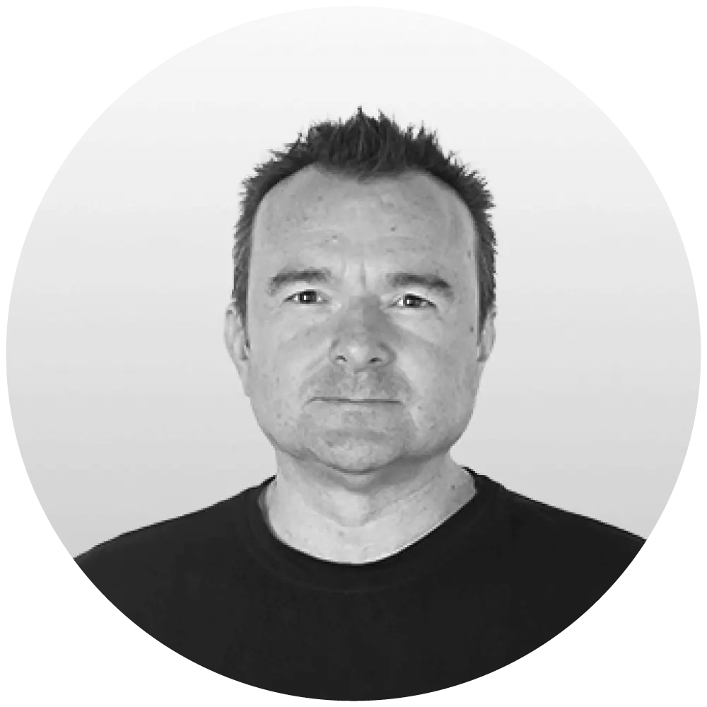 A black and white headshot of a person with short hair, looking directly at the camera. They have a neutral expression, wearing a dark shirt