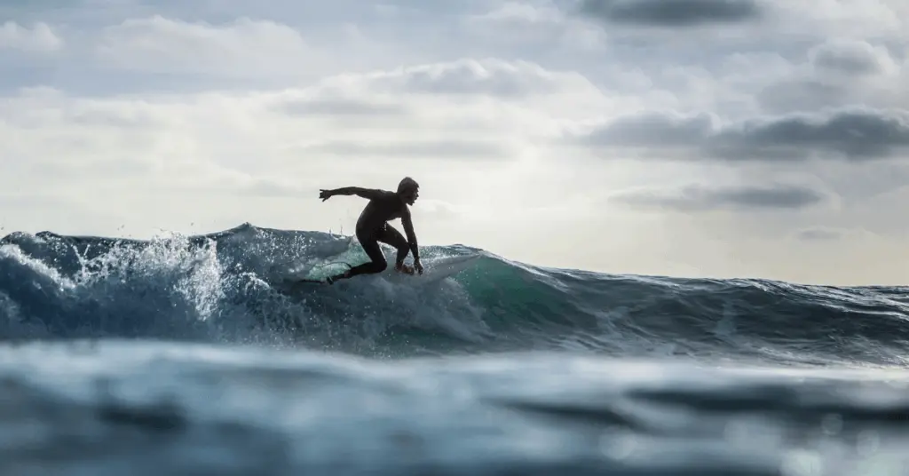 Surfer in wetsuit gracefully rides a breaking wave under a cloudy sky. The scene conveys a sense of adventure and freedom in the ocean